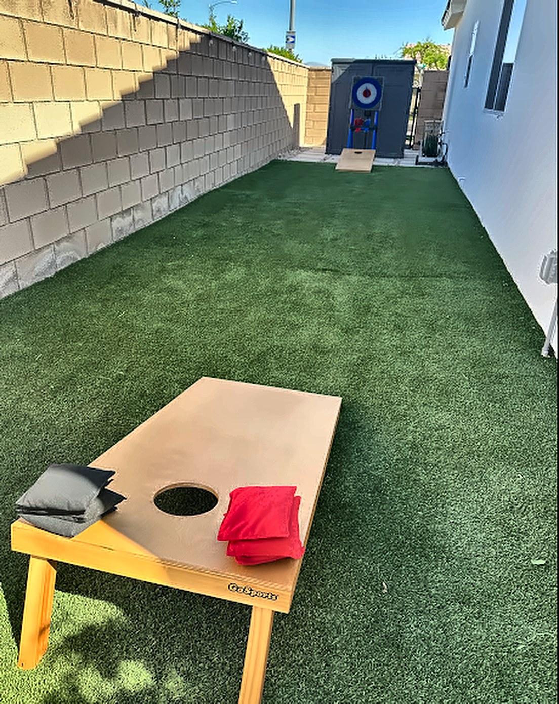 Cornhole board with red and black bags on the turf side yard, with the axe-throwing target at the far end