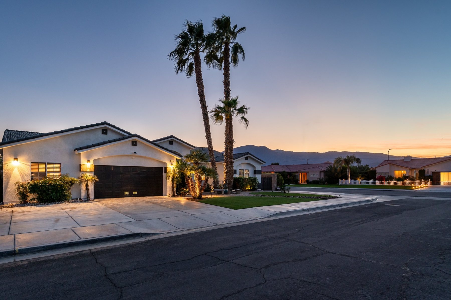 Front of the house at dusk with landscape lighting, three tall palms against the sunset, and the main driveway